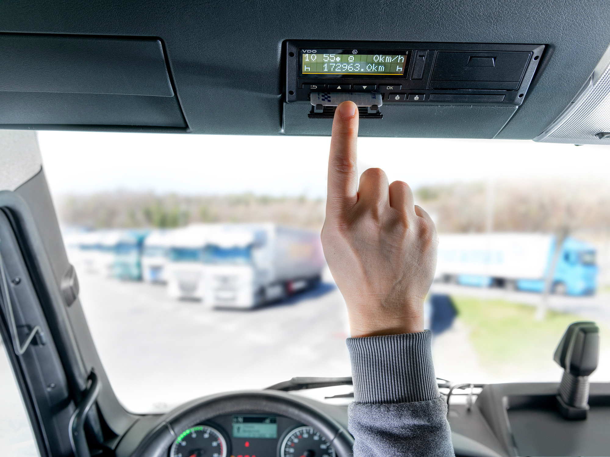 A driver inserts his driver card into the VDO DCTO 4.1 - the second version of the intelligent tachograph. Other trucks in the fleet can be seen blurred in the background through the windshield.